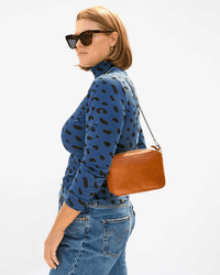 A woman in dark sunglasses, a blue spotted long-sleeve shirt, and blue jeans stands sideways with the Clare V. Midi Sac in Tan Napetto on her shoulder against a plain light background.