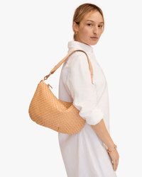 A woman in a white button-down dress stands sideways, looking at the camera, carrying the Clare V. Moyen Messenger in Bisque Woven Checker. She wears minimal jewelry—hoop earrings and a bracelet—against a plain white background.
