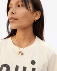 A woman with long dark hair and freckles wears a cream shirt and vintage gold jewelry, including hoop earrings and two necklaces—a heart pendant and the Clare V. Padlock Locket in Vintage Gold. She gazes slightly to the left.