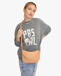 A person with light brown hair, wearing a grey “PAS MAL” sweatshirt, a necklace, a ripped denim skirt, and carrying the Clare V. Petit Moyen Messenger in Bisque Woven Checker stands with one hand on the bag strap.