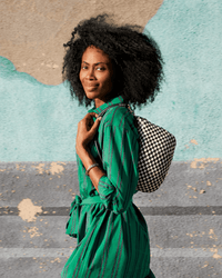 A woman in a green striped dress smiles while walking, carrying the Clare V. Woven Checker Petit Moyen Messenger in Black & Cream over her shoulder, standing against a colorful abstract wall.