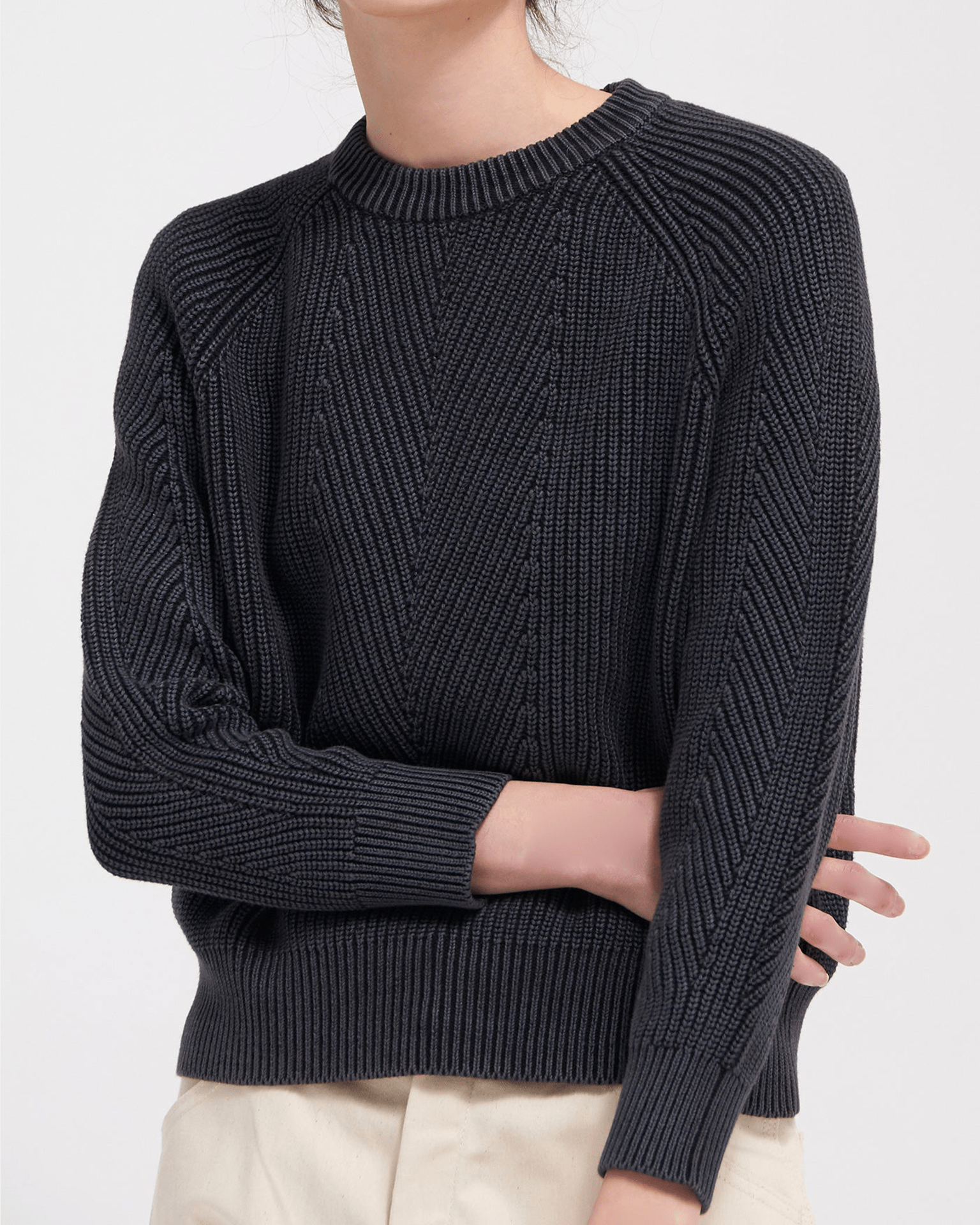 A person crosses one arm over their body against a plain background, wearing Demylee’s Chelsea Washed Cotton Sweater in Black—a dark, textured pullover with ribbed collar, cuffs, hem—and light-colored pants.