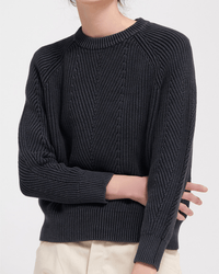 A person crosses one arm over their body against a plain background, wearing Demylee’s Chelsea Washed Cotton Sweater in Black—a dark, textured pullover with ribbed collar, cuffs, hem—and light-colored pants.