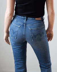 A person wearing a black sleeveless top and Edwin's Lark Ankle in Geary jeans stands with their back to the camera, showcasing the jeans' waistband and back pockets.