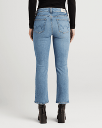 A person is standing with their back to the camera against a white background, wearing Edwin's Lark Ankle in Lombard jeans, black heeled boots, and a black long-sleeve top.