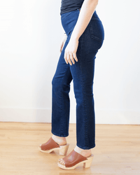 Someone stands indoors on a wooden floor against a white wall, shown from the side, wearing Edwin’s Lark Ankle Pull-On in Quietude jeans, paired with a black top and brown heeled sandals.