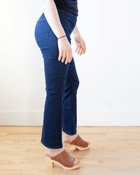 A person in Edwin's Lark Ankle Pull-On jeans in Quietude and a black top with brown heeled sandals stands on a light wood floor against a plain white wall, their body angled slightly to the side.