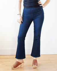 Someone wearing Edwin's Lark Ankle Pull-On in Quietude jeans, a black sleeveless top, and brown heeled sandals stands on a wooden floor against a light-colored wall. Their face is not visible.