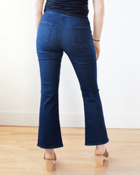 A person stands indoors on a wooden floor, facing away with relaxed hands, wearing Edwin's Lark Ankle Pull-On in Quietude jeans and tan heeled sandals.