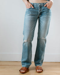 Person standing indoors wearing Edwin's Simone in Jet Stream, medium indigo, slightly distressed midrise straight leg jeans with brown sandals and hands in front pockets, shown from waist to feet.