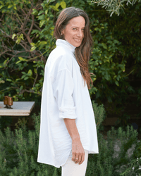A person with long hair stands outdoors, smiling slightly against a green backdrop, wearing the Shirley Oversized Button-Up Shirt in White Distressed Famous Denim by Frank & Eileen.