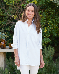 A woman wearing a Frank & Eileen Shirley Oversized Button-Up Shirt in White Distressed Famous Denim smiles while standing in a garden with lush greenery around her.