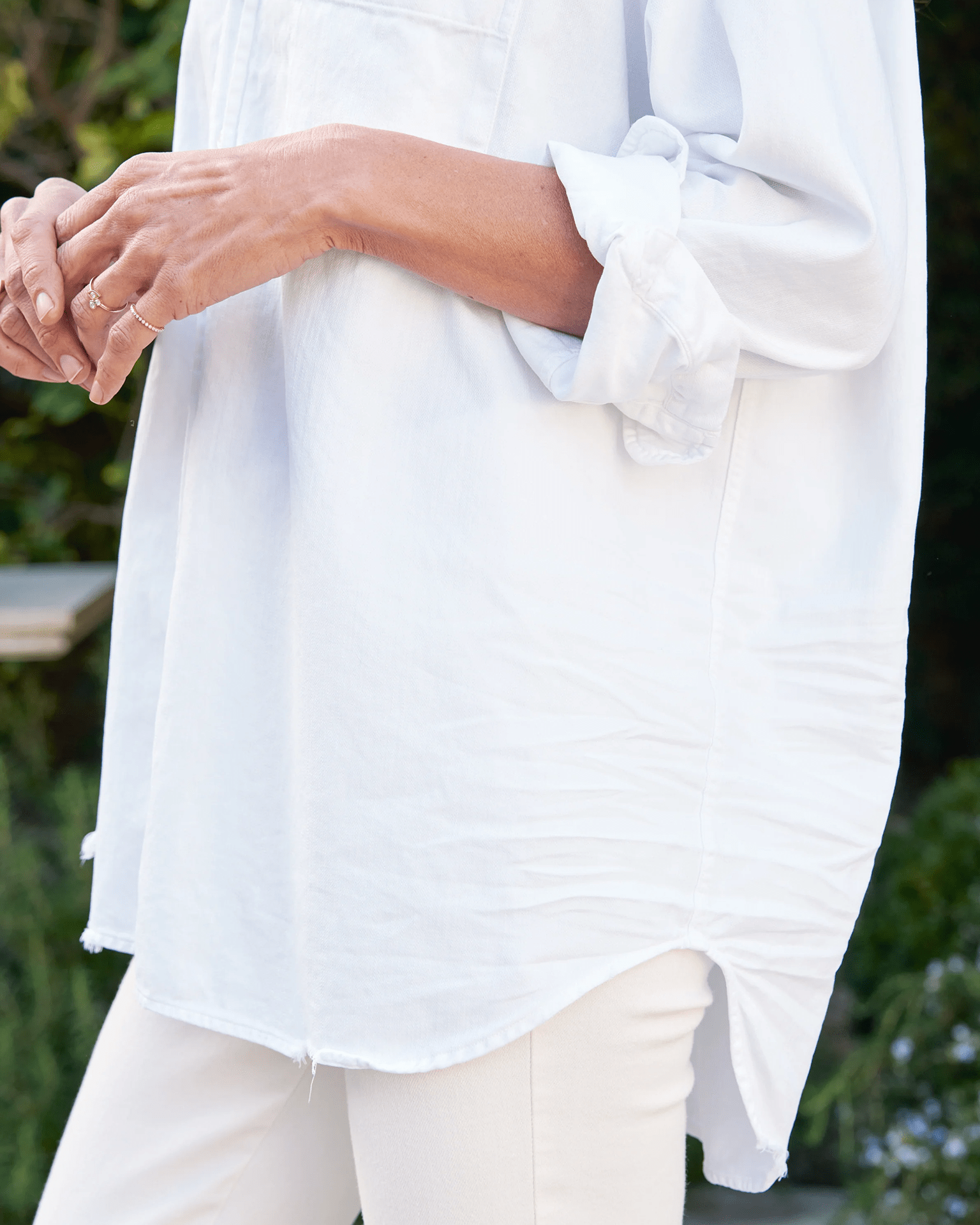 A person wearing the Shirley Oversized Button-Up Shirt in White Distressed Famous Denim by Frank & Eileen and white pants stands outdoors, greenery in the background, with their hands slightly clasped.