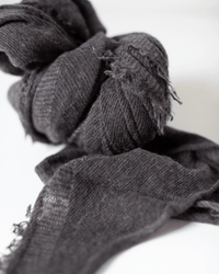 A close-up of Grisal's Love Cashmere Scarf in Brown Top X Charcoal, knotted loosely on a white surface. The dark gray tones and frayed edges reveal its heirloom quality and lasting elegance.