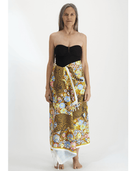 A woman with long gray hair stands facing forward in a black strapless top and a colorful Inoui Editions sarong, against a white background. She has the Inoui Editions Fouta 100 Pampa towel in Caramel draped casually over her arm.