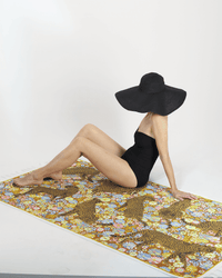 A person in a black swimsuit and wide-brimmed hat sits on the Inoui Editions Fouta 100 Pampa beach towel in Caramel, featuring bold patterns, against a plain white background.