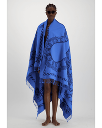 A barefoot person in sunglasses is wrapped in the Fouta 140 3D Mantra in Blue by Inoui Editions, featuring dark abstract patterns and fringe, set against a plain light gray background.