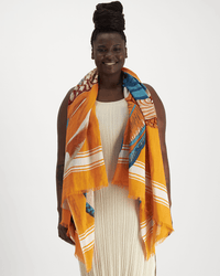 A smiling woman with dark braided hair in a bun wears a cream dress and the Inoui Editions Scarf 100 Toucan in Orange, featuring vibrant orange, blue, and white tones, draped over her shoulders against a plain white background.