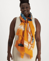 A person in a sleeveless cream knit dress and an Inoui Editions Scarf 100 Toucan in Orange smiles against a plain white background.