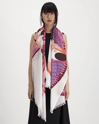 A woman with straight black hair and bangs stands against a white background, wearing a long black dress and an Inoui Editions Scarf 100 Toucan in Pink with pink, orange, purple, and white patterns draped over her shoulders.