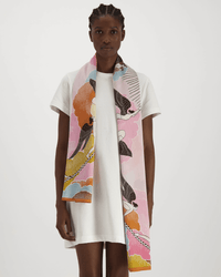 A person in a plain white t-shirt dress stands facing forward with the Inoui Editions Scarf 100 Yoga in Yellow, featuring birds, clouds, and abstract shapes in pink, orange, yellow, black, and white, draped around their neck.