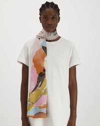 A person in a plain white short-sleeve dress stands against a white background, wearing the Inoui Editions Scarf 100 Yoga in Yellow—a cotton silk scarf with abstract pink, orange, yellow, and black patterns—draped around their neck.