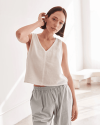 A person with short brown hair wears the It is well LA Sleeveless Crop Top in Salt, paired with loose light gray pants, standing indoors in soft natural light and touching the side of their head.
