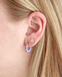 A close-up of an ear with a small round silver stud in the upper lobe and Kris Nations Twisted Enamel Hoop Earrings in Peri—lavender enamel coated brass with gold accents—in the lower lobe. The person has light blonde hair.
