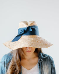 A woman with long brown hair models the Lola Hats First Aid in Natural/Cadet, a wide-brimmed summer hat with a large navy ribbon, partially covering her face. She wears a denim shirt over a white top against a plain light background.
