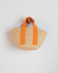 The Lola Hats Mini Market Bag in Cream/Orange features two bold orange handles and hangs on a wooden hook against a white wall—a chic, handmade raffia accessory ideal for summer outings.