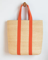 The Lola Hats PB & J Bag w/ Long Handle in Cream/Carrot, a woven raffia tote with bold orange handles, hangs on a peg against a white wall—a perfect handmade summer accessory from Brooklyn.