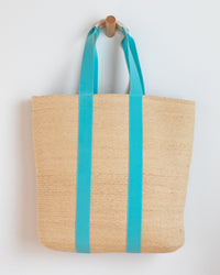 The Lola Hats PB & J Bag w/ Long Handle in Cream/Turquoise, a woven straw tote with bright turquoise handles, hangs on a wooden peg against a white wall.