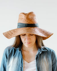 A woman wearing the Snap First Aid hat in Caramel/Pewter by Lola Hats and a denim shirt looks down with a neutral or slightly sad expression against a plain white background.