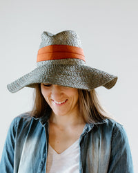 A woman with long brown hair smiles while looking down, wearing Lola Hats' Snap First Aid in Sage/Rust—a packable raffia summer hat with an orange band—paired with a light denim shirt and white top against a plain white background.