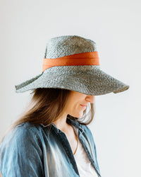 A woman in profile wears the Lola Hats Snap First Aid in Sage/Rust, a wide-brimmed packable raffia hat with an orange band. She has long brown hair, a blue denim shirt over a white top, and stands against a plain light background.