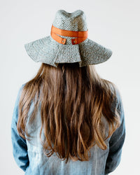 A person with long brown hair, dressed in a light blue denim shirt and wearing the Lola Hats Snap First Aid in Sage/Rust, is seen from behind against a plain white background.