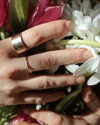 A close-up of a hand wearing the Nashelle Pure Ring in Gold gently holds a bouquet of pink and white flowers in bright natural light.