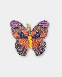 The Butterfly Brooch in Purple by Olivia Dar features detailed, handwoven wings and body crafted from orange, purple, yellow, and dark blue beads on a white background.