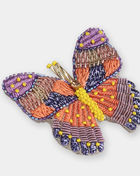 The Butterfly Brooch in Purple by Olivia Dar is handwoven with orange, purple, and yellow beads and threads, featuring intricate patterns and textured wing and body details.