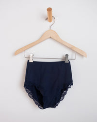 The Del w/ Lace High Cut Brief in Navy by Only Hearts hangs on a wooden hanger against a white wall.
