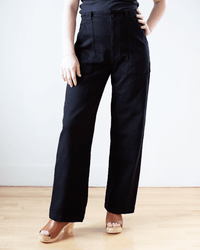 A person stands indoors wearing Prairie Underground's Long Carpenter Pant in Black with a black top and tan platform sandals. Only the lower body is shown, one hand on the hip, highlighting the pant's high-rise and wide-leg silhouette.