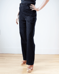 Someone wearing a black t-shirt, Prairie Underground Long Carpenter Pant in Black, and light-colored platform sandals stands against a plain white wall on a wooden floor.