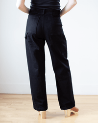 A person wearing Prairie Underground's Long Carpenter Pant in Black stands indoors on a light wooden floor, facing away from the camera against a plain white wall.