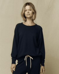 A person with short, light brown hair wears the Great's The College Sweatshirt in True Navy and matching pants with a white drawstring, standing against a plain beige background.