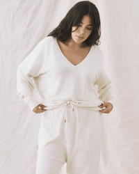 A woman with long dark hair wears the Great's The V-Neck Sweatshirt in Washed White and matching drawstring pants, standing against a light fabric backdrop with hands in her pockets, looking down.