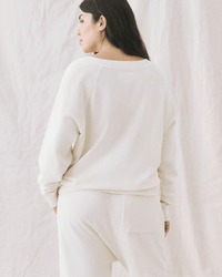 A person with long dark hair stands facing away, wearing the Great’s The V-Neck Sweatshirt in Washed White and matching pants against a soft, neutral fabric backdrop.