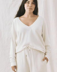 A woman with long dark hair wears the Great’s V-Neck Sweatshirt in Washed White and matching drawstring pants, both in a relaxed, boxy fit, standing against a soft light-colored fabric backdrop with one hand in her pocket.