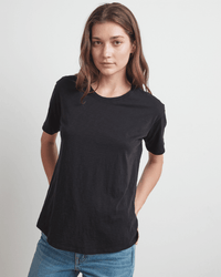 A woman with long brown hair wears the Velvet by Graham & Spencer Tera S/S Tee in Black and blue jeans, standing against a white background. She looks at the camera with a neutral expression, her hands behind her back.