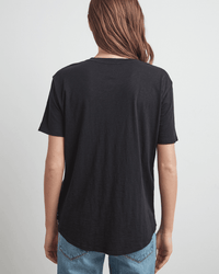 A person with long brown hair is shown from behind, wearing the Tera S/S Tee in Black by Velvet by Graham & Spencer and blue jeans, standing against a light gray background.