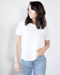 A woman with wavy dark hair, wearing the Tera S/S Tee in White by Velvet by Graham & Spencer and light blue jeans, stands against a plain light background, looking to her left with one hand in her pocket.